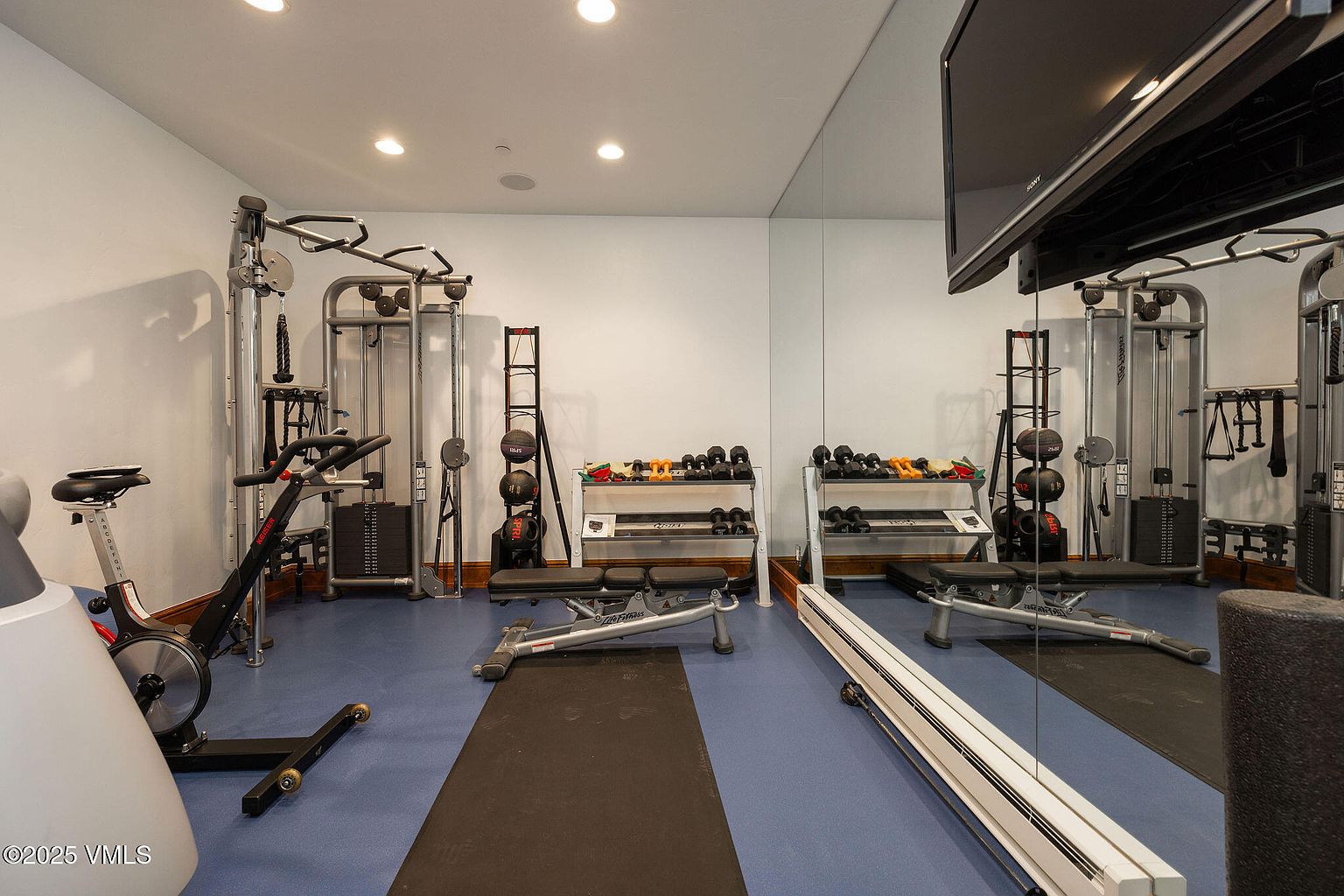 This is an interior shot of a well-equipped home gym. The room features various exercise machines, including a stationary bike and weightlifting stations, along with dumbbells and medicine balls. A large mirror covers one wall, enhancing the sense of space and allowing for form checks during workouts.