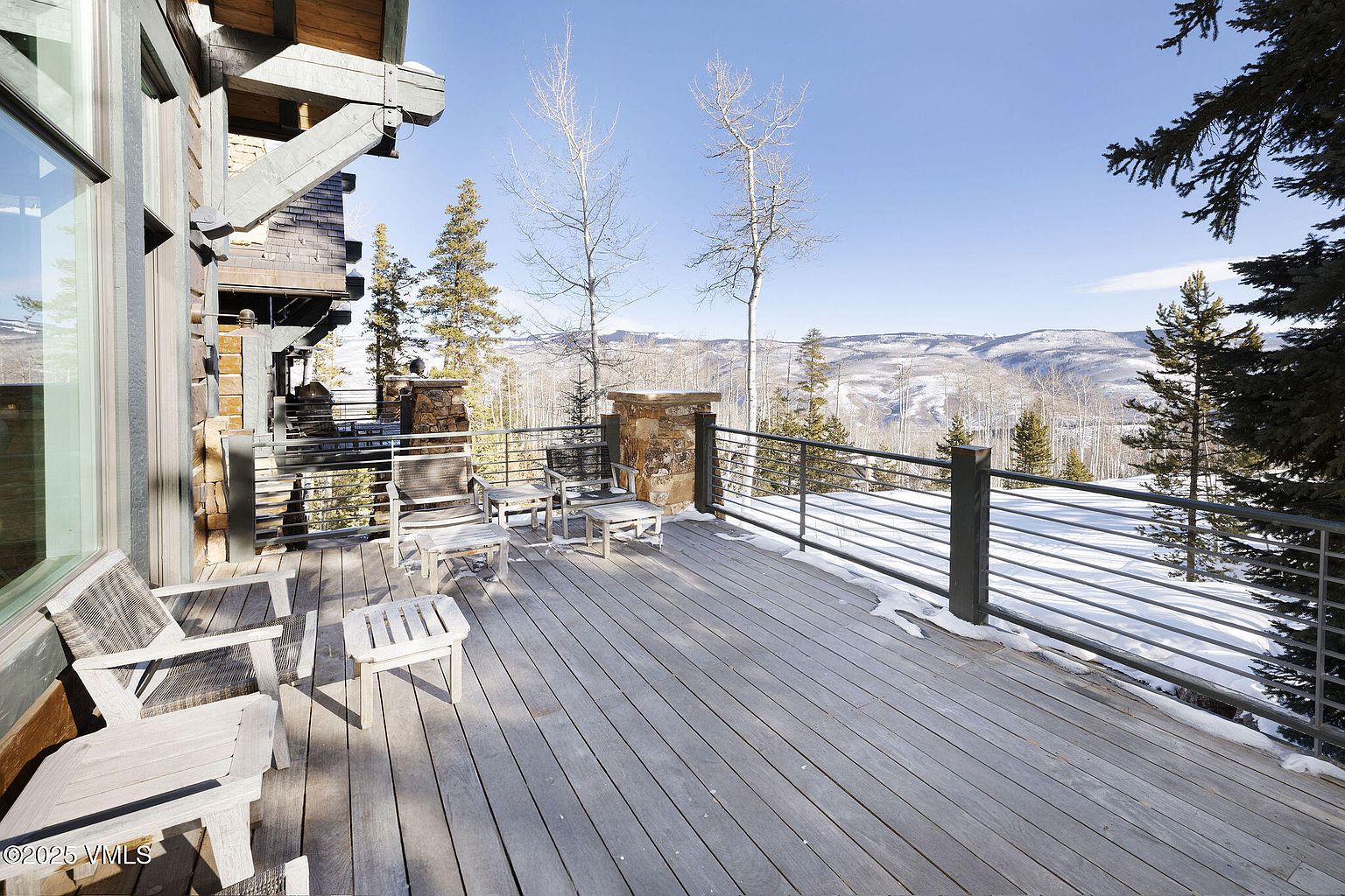 This image showcases a spacious wooden deck with outdoor seating, offering a serene view of snow-covered mountains and trees. The deck features a metal railing and stone pillars, blending rustic charm with modern design. The scene evokes a sense of tranquility and connection with nature, ideal for relaxation and outdoor enjoyment.