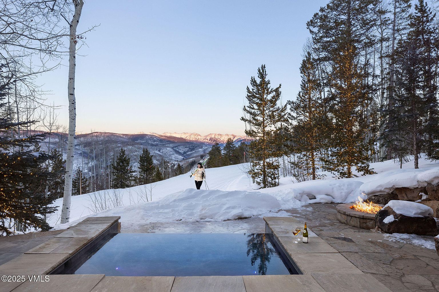This image showcases an inviting outdoor spa area with a rectangular pool reflecting the sky, set against a snowy mountain backdrop. A stone fire pit with flames adds warmth and ambiance, while a bottle of wine and glasses suggest relaxation. The scene is framed by snow-covered trees, some adorned with lights, creating a luxurious and serene winter retreat.