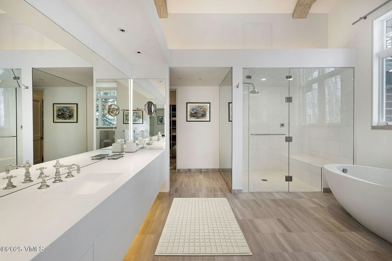 This is an elegantly designed primary bathroom, featuring a long, white countertop with dual sinks and stylish faucets. A glass-enclosed shower and a modern soaking tub provide luxurious bathing options. The neutral color palette creates a serene and spa-like atmosphere, complemented by wood-look flooring.
