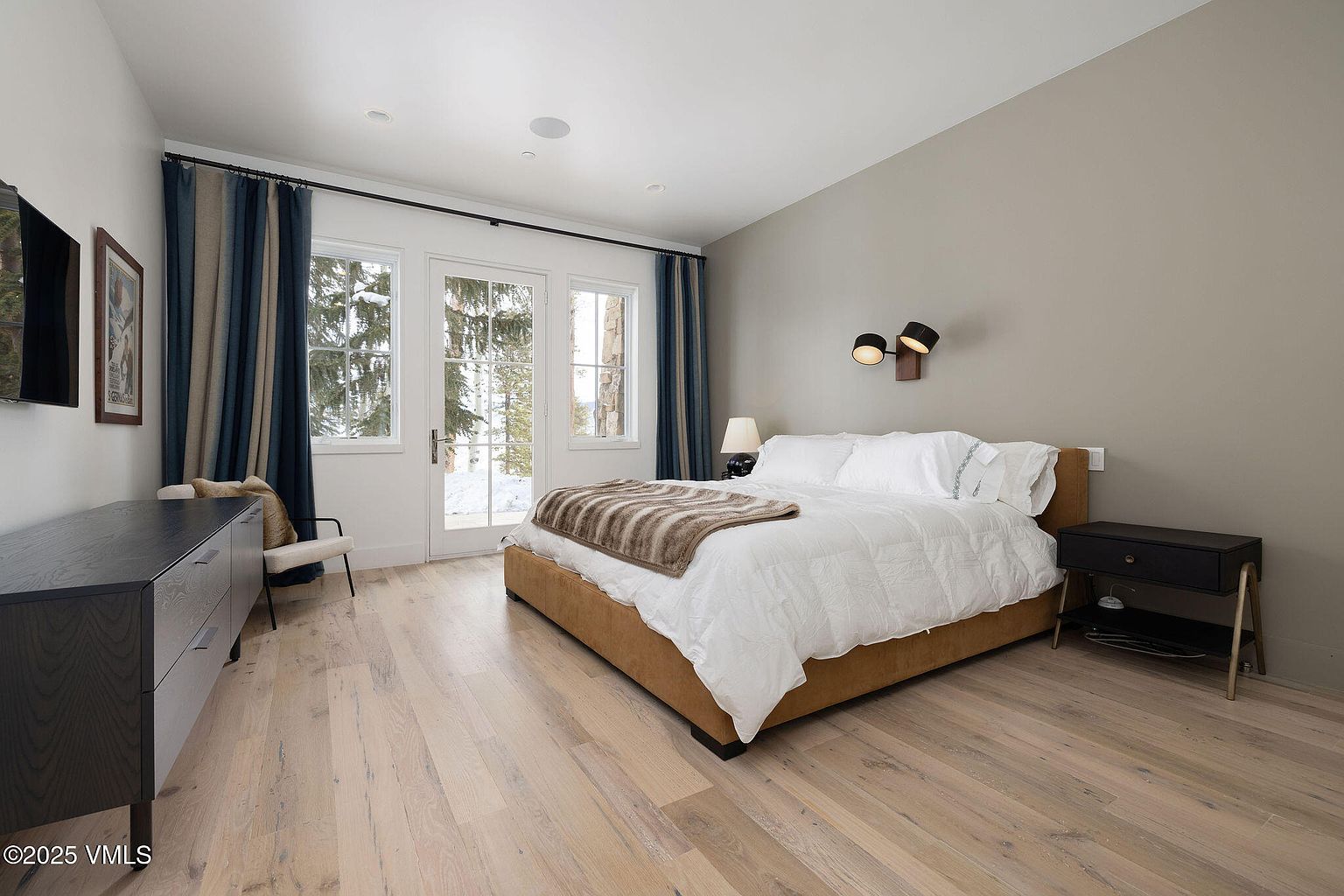 This is a primary bedroom featuring a king-size bed with a tan upholstered frame, white bedding, and a striped throw blanket. The room has light hardwood floors, a dark wood dresser, and a nightstand. Natural light enters through the French doors, which offer a view of a snowy landscape.