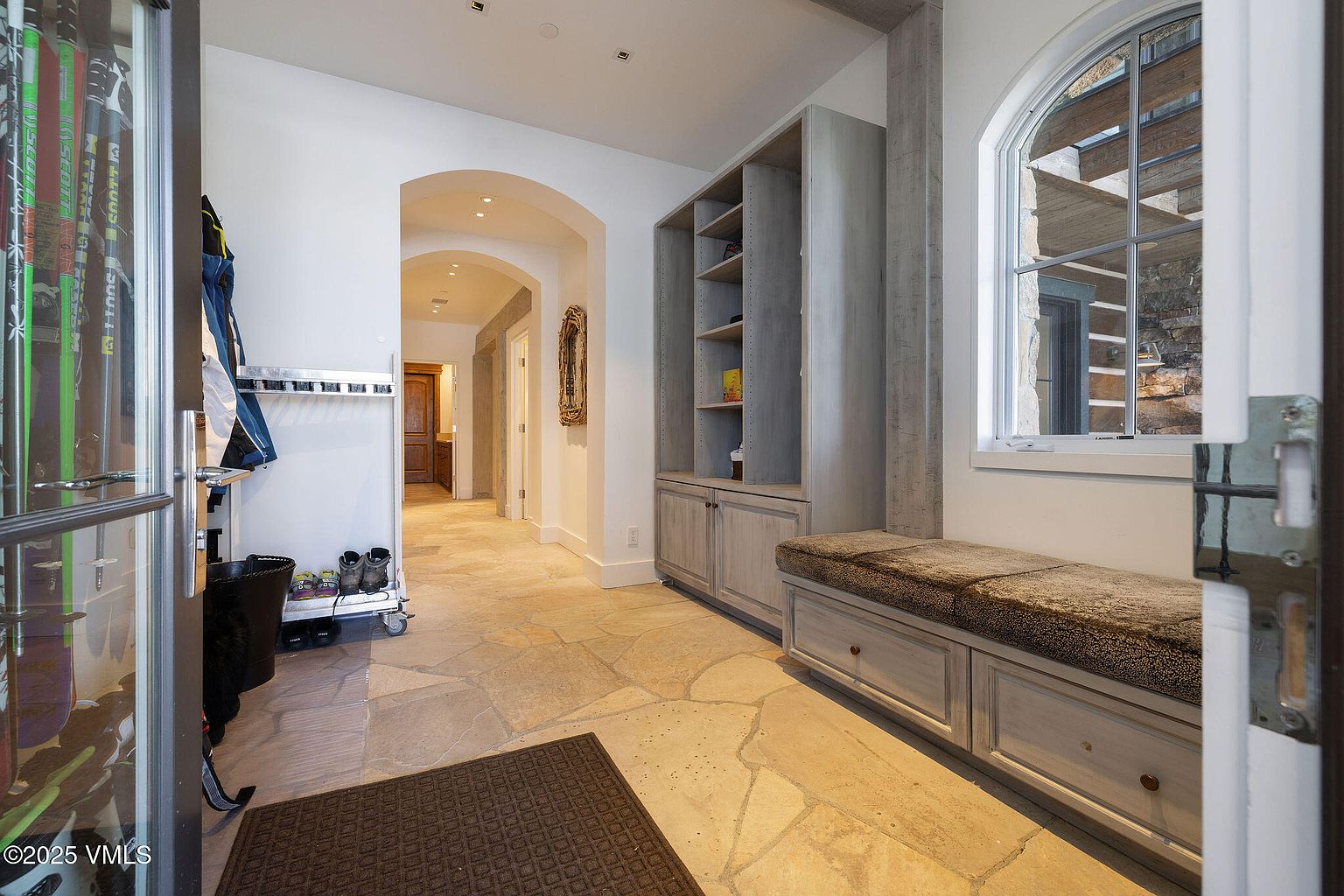 This interior shot showcases a hallway or entryway featuring stone flooring and white walls. A built-in storage unit with shelving and a cushioned bench seat is visible on the right. The space is well-lit, leading to an arched doorway further down the hall, creating a welcoming and functional transition area.