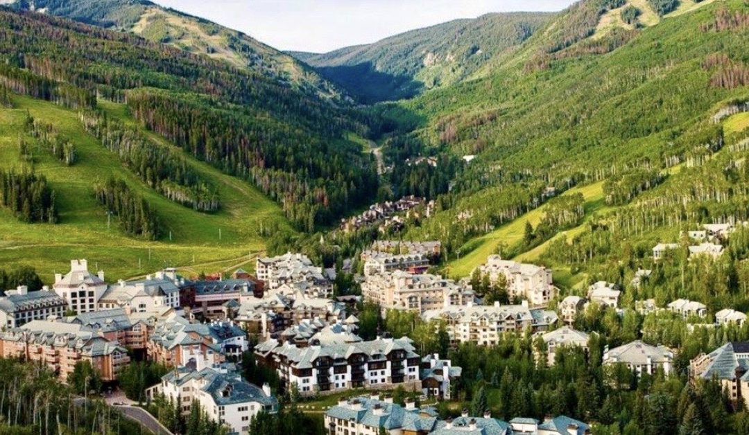 This aerial view showcases a luxurious residential area nestled within a lush valley. The architecture features a blend of grand homes and lodges, characterized by their multi-story designs and varied rooflines. Surrounded by dense forests and rolling green hills, the setting offers privacy and scenic beauty, making it an attractive real estate opportunity for those seeking a serene retreat.