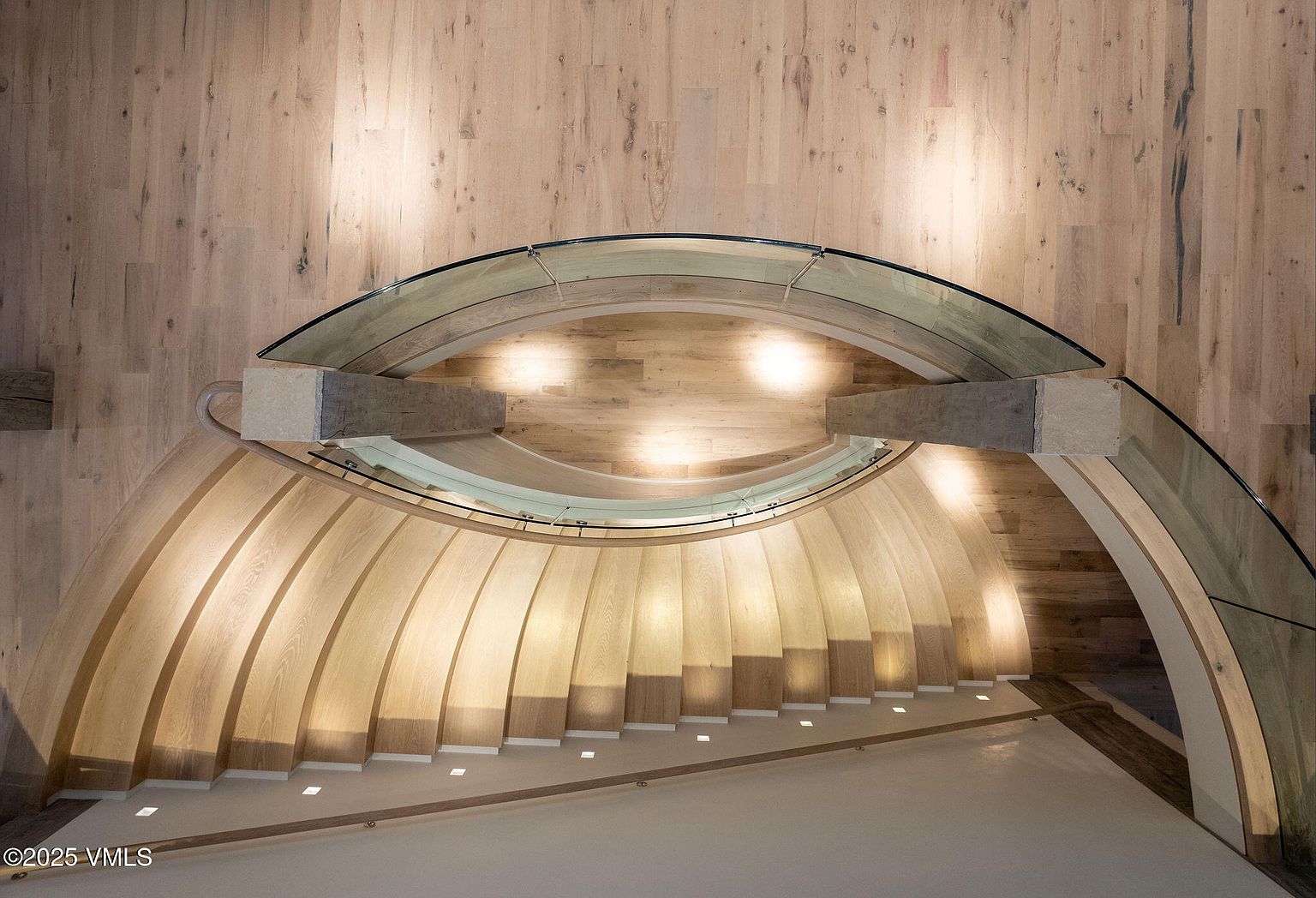 This image showcases a modern, curved staircase with wooden steps and a glass railing. The staircase is illuminated by subtle lighting embedded in the steps, creating a warm and inviting ambiance. The surrounding walls and flooring are also wood, contributing to a cohesive and luxurious design.