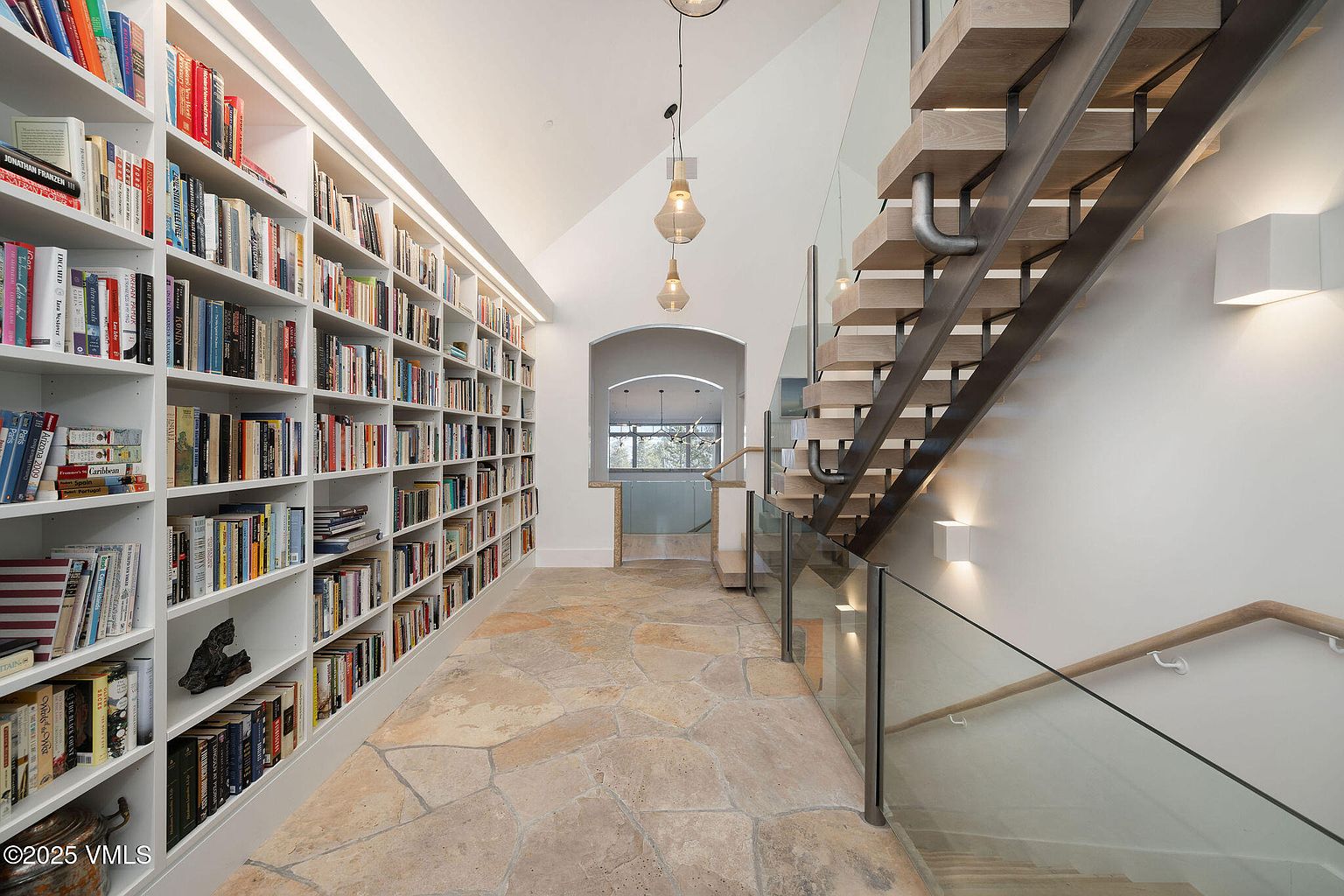 This interior shot showcases a hallway with a striking combination of design elements. A floor-to-ceiling bookshelf lines one wall, filled with books, while a modern staircase with glass railings ascends on the opposite side. The flooring is a natural stone, and pendant lights hang from the ceiling, adding to the sophisticated ambiance.