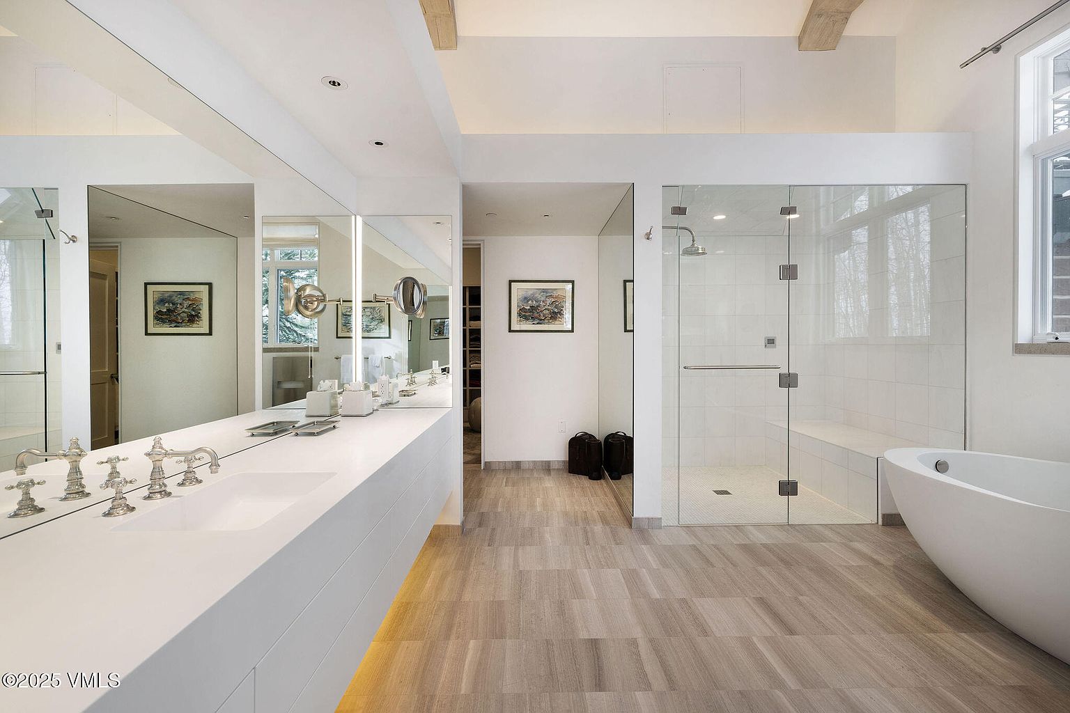 This is a modern primary bathroom featuring a long, sleek white countertop with integrated sinks and chrome fixtures. Large mirrors extend across the wall above the countertop, reflecting the light and creating a sense of spaciousness. A glass-enclosed shower with a built-in bench and a freestanding bathtub are also visible, contributing to the bathroom's luxurious and contemporary design.