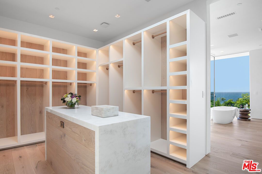 This luxurious, modern walk-in closet features custom light-wood cabinetry with integrated LED lighting and a central island topped with white marble. The open-concept design flows seamlessly into an adjacent bathroom, which offers a stunning, unobstructed view of the ocean through floor-to-ceiling glass. The space exudes a high-end, serene atmosphere with its clean lines and sophisticated material palette.
