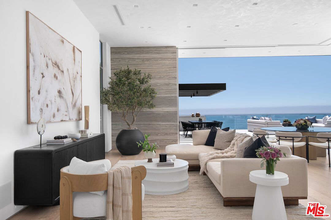This sophisticated, open-concept living room features a neutral color palette, plush cream-colored sectional seating, and a modern wooden armchair. The space is anchored by a textured area rug and a minimalist white coffee table, while a large potted olive tree adds a touch of organic elegance against a striking stone accent wall. Large floor-to-ceiling glass doors seamlessly blend the interior with an expansive outdoor terrace, offering a breathtaking, unobstructed view of the ocean.