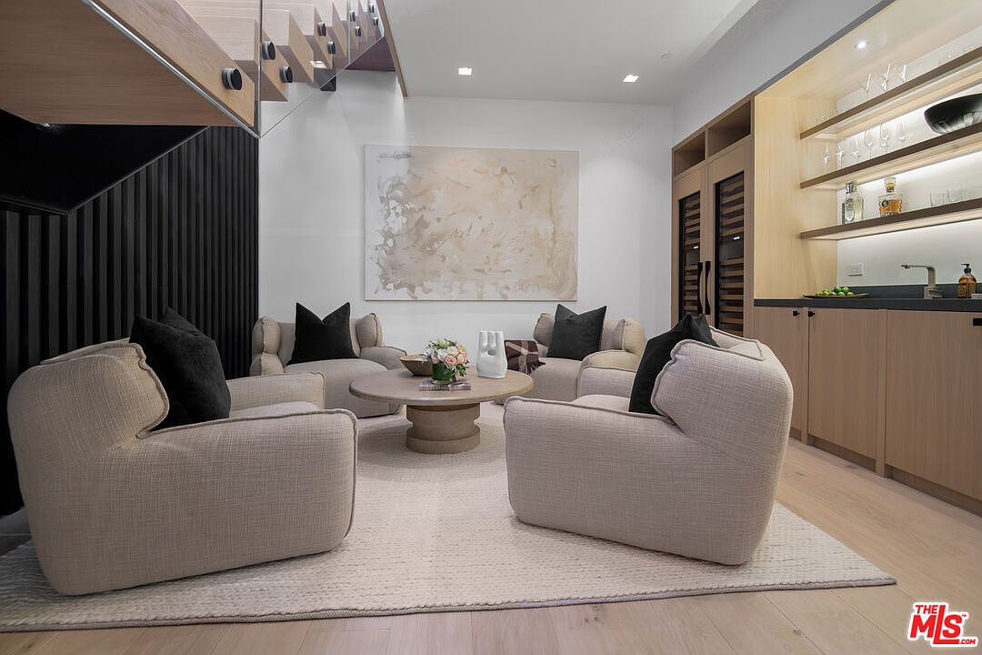This sophisticated lounge area features a symmetrical arrangement of four plush, light-toned armchairs centered around a round wooden coffee table on a textured area rug. The space is accented by a large abstract painting, a sleek built-in wet bar with glass shelving, and a modern staircase with glass railings visible on the left. The overall aesthetic is contemporary and luxurious, characterized by a neutral color palette and clean, minimalist lines.
