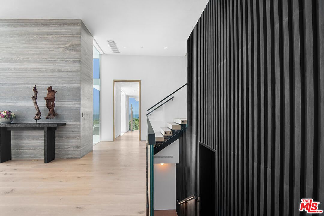 This sophisticated interior space features a striking contrast between a textured, grey-toned stone wall and a dramatic, floor-to-ceiling black slatted wood accent wall. A modern staircase with glass railings and floating wooden treads leads downward, while a bright, open hallway extends toward a distant view of the outdoors. The minimalist design, combined with high-end materials and clean lines, creates an atmosphere of luxury and architectural precision.