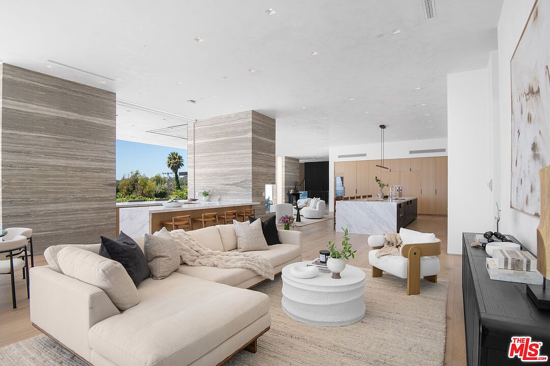 This expansive, open-concept living area showcases a sophisticated modern aesthetic with high ceilings, light wood flooring, and striking floor-to-ceiling stone accent walls. The space is anchored by a plush, cream-colored sectional sofa and a textured white coffee table, seamlessly flowing into a sleek kitchen with a marble-topped island in the background. Large glass walls provide a bright, airy atmosphere and a clear view of the lush outdoor greenery, creating a luxurious and seamless indoor-outdoor living experience.