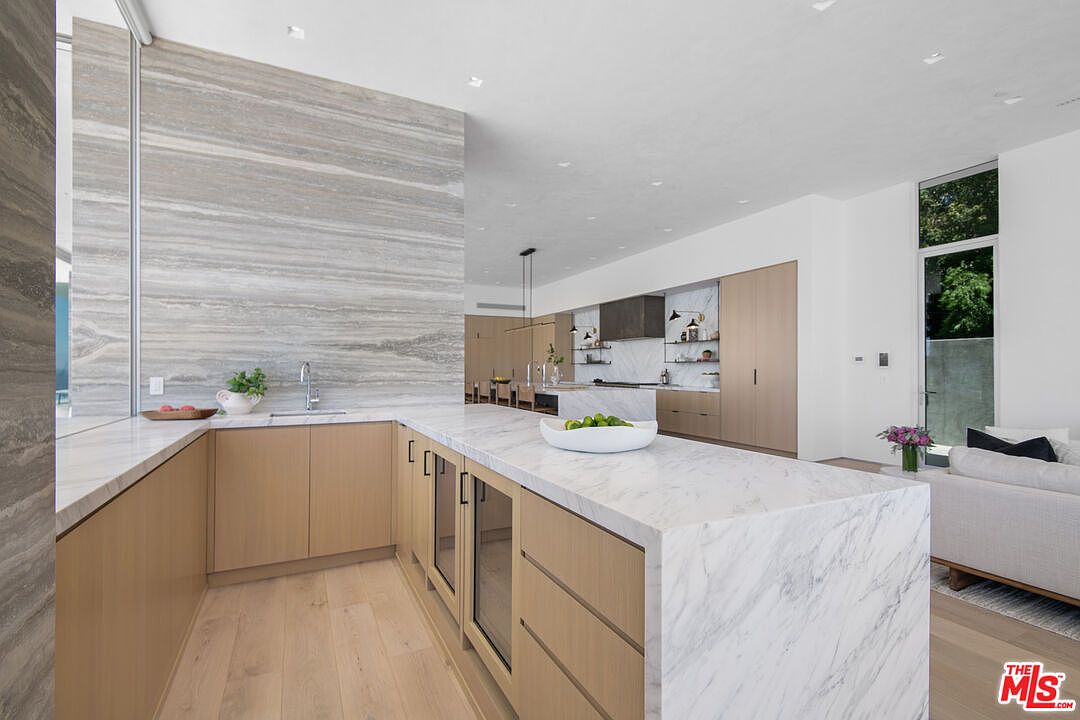 This ultra-modern, high-end kitchen features a seamless blend of light wood cabinetry and striking marble countertops that extend into a large waterfall island. The space is defined by a dramatic, floor-to-ceiling stone accent wall and clean, minimalist lines that create an airy, sophisticated atmosphere. The perspective captures the expansive layout, highlighting the integration of the kitchen with the adjacent living area and the bright, natural light streaming in from the large windows.