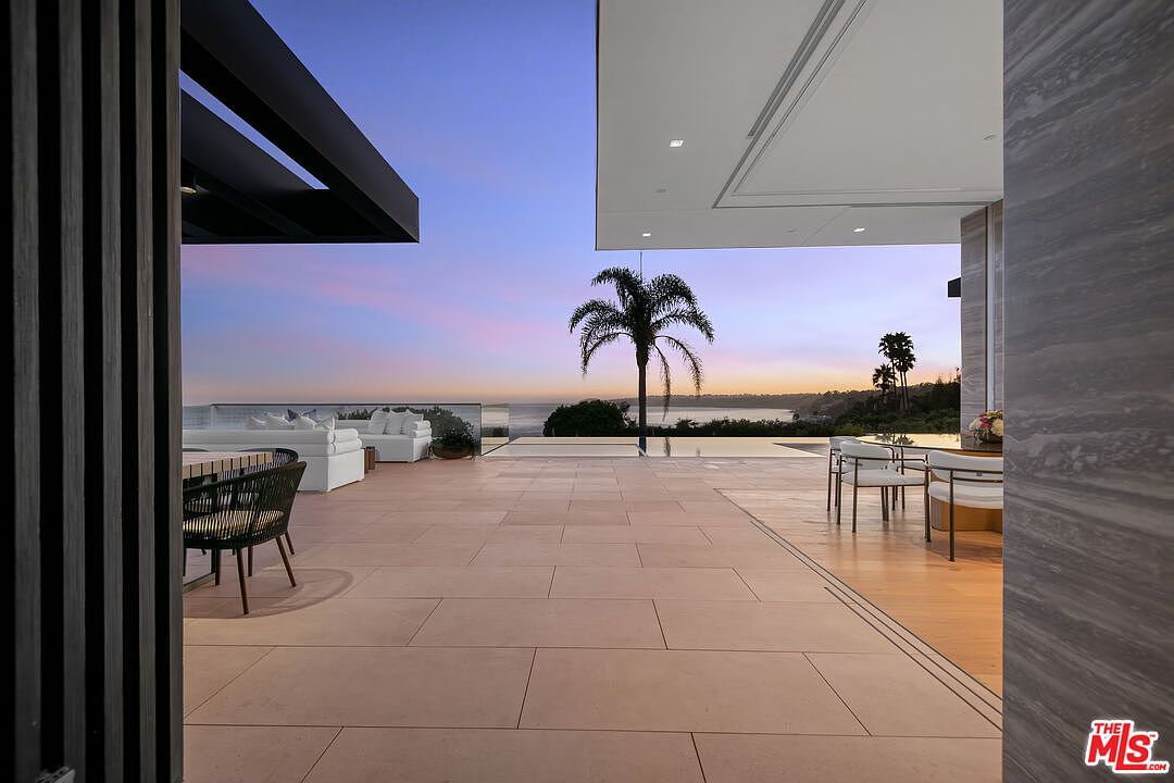This luxurious, expansive outdoor patio seamlessly blends indoor and outdoor living with its open-concept design and large sliding glass doors. The space features high-end modern furniture, a sleek infinity-edge pool, and a breathtaking panoramic view of the ocean at sunset. The clean lines, neutral stone flooring, and minimalist architecture create a sophisticated and serene atmosphere perfect for high-end entertaining.