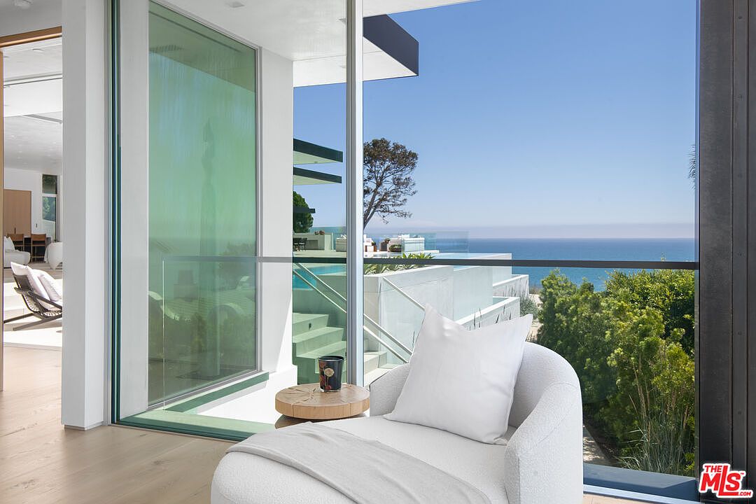 This interior shot captures a serene lounge area featuring a plush, white curved armchair positioned next to a small, natural wood side table. The space is defined by floor-to-ceiling glass walls that seamlessly blend the indoor living area with a stunning outdoor balcony, infinity pool, and expansive ocean views. The minimalist, modern aesthetic emphasizes light, luxury, and a strong connection to the coastal environment.