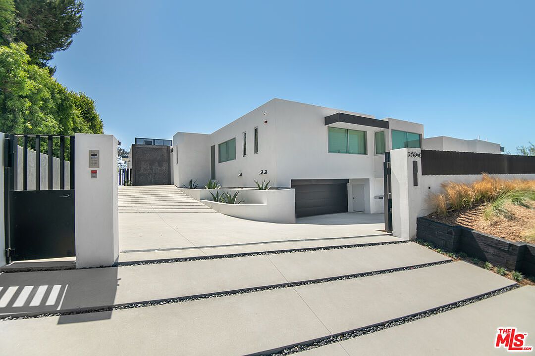 This modern, minimalist residence features a striking white stucco facade with clean lines and large, rectangular windows. The property is accessed via a wide concrete driveway leading to an integrated garage, framed by a sleek black metal gate and contemporary landscaping. The overall aesthetic is sophisticated and architectural, emphasizing a high-end, private suburban lifestyle.