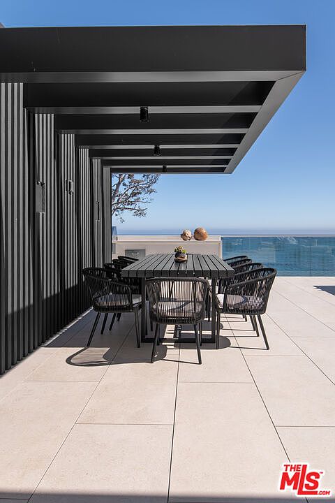 This sleek, modern outdoor dining area features a minimalist black pergola structure extending over a contemporary dining set with woven chairs. The space is set on a large-format tiled deck that overlooks a stunning, unobstructed ocean view, creating a sophisticated and serene atmosphere. The clean lines and monochromatic color palette emphasize a high-end, architectural aesthetic perfect for luxury coastal living.