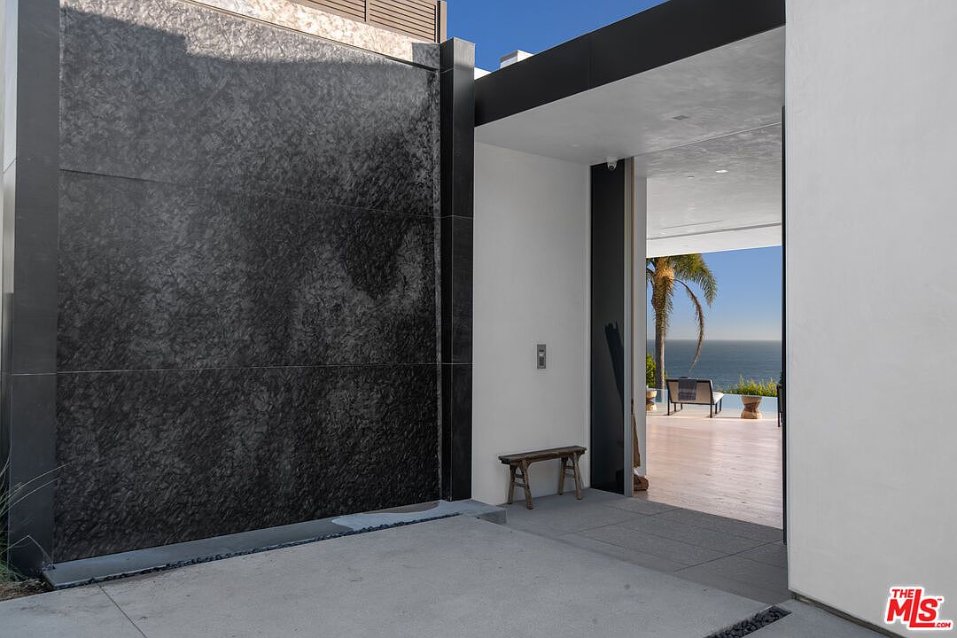 This striking entryway features a dramatic, textured black stone water wall that contrasts beautifully with the clean, minimalist white architecture. The open doorway frames a breathtaking view of the ocean and a sun-drenched patio, creating a seamless transition between the home's interior and the coastal landscape. A rustic wooden bench sits near the entrance, adding a touch of organic warmth to the sophisticated, modern design.