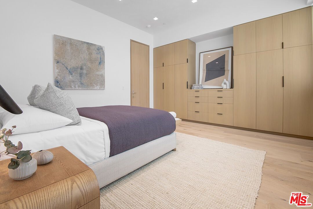 This modern primary bedroom features a minimalist aesthetic with light wood flooring, a neutral color palette, and a large built-in wardrobe system. A plush bed with a dark purple throw blanket serves as the focal point, complemented by a textured area rug and a sleek wooden nightstand. The room is bright and airy, highlighted by recessed lighting and a large abstract piece of art on the wall.