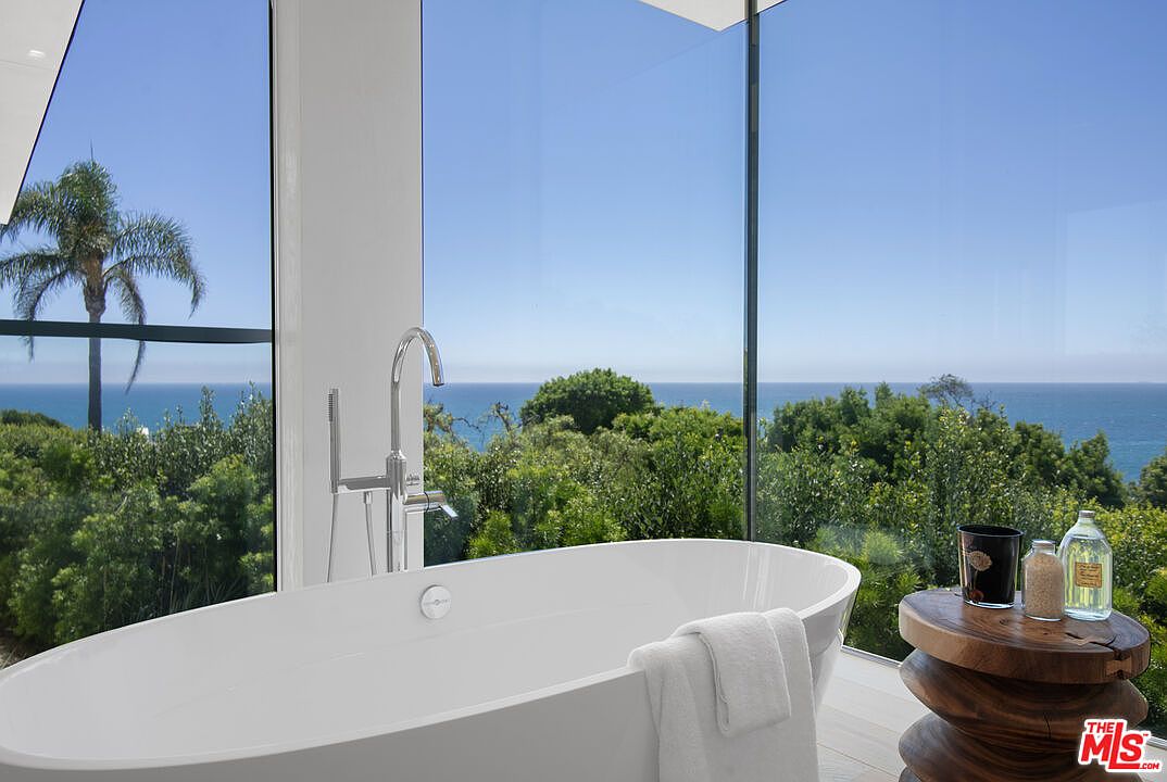 This luxurious primary bathroom features a sleek, freestanding white soaking tub positioned perfectly against floor-to-ceiling glass windows that offer a breathtaking, unobstructed view of the ocean and lush greenery. A rustic, natural wood side table holds bath essentials, adding a warm, organic contrast to the modern, minimalist aesthetic. The perspective is intimate and serene, emphasizing the seamless integration of high-end interior design with the natural beauty of the coastal landscape.