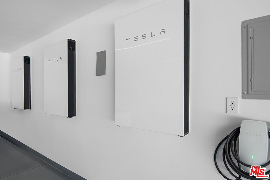 This image showcases a clean, modern garage wall equipped with three Tesla Powerwall battery units and a Tesla Wall Connector charging station. The minimalist white walls and organized setup highlight the home's advanced energy storage and electric vehicle infrastructure. The perspective is a wide-angle shot looking down the side wall, emphasizing the sleek, high-tech aesthetic of the space.