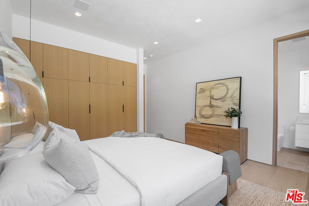 This modern primary bedroom features a minimalist aesthetic with light wood flooring, a large built-in wardrobe, and a neutral color palette. A plush bed with white linens sits in the foreground, while a wooden dresser topped with abstract art and a small vase adds warmth to the space. The room flows seamlessly into an adjacent bathroom, creating an open and airy atmosphere.
