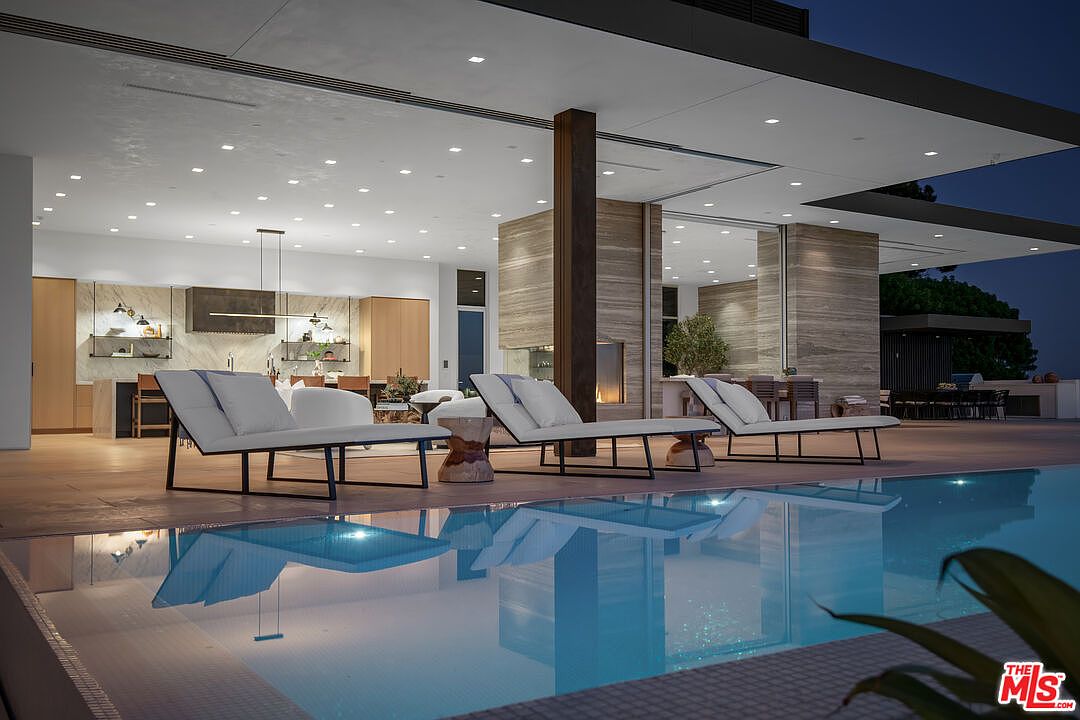 This stunning contemporary outdoor living space features a sleek, illuminated infinity-edge pool that seamlessly blends with the indoor-outdoor transition of the home. The patio is furnished with modern lounge chairs, while the interior reveals a high-end kitchen and living area, all unified by warm, natural stone textures and recessed lighting. The perspective captures a sophisticated, resort-like atmosphere perfect for evening entertaining.