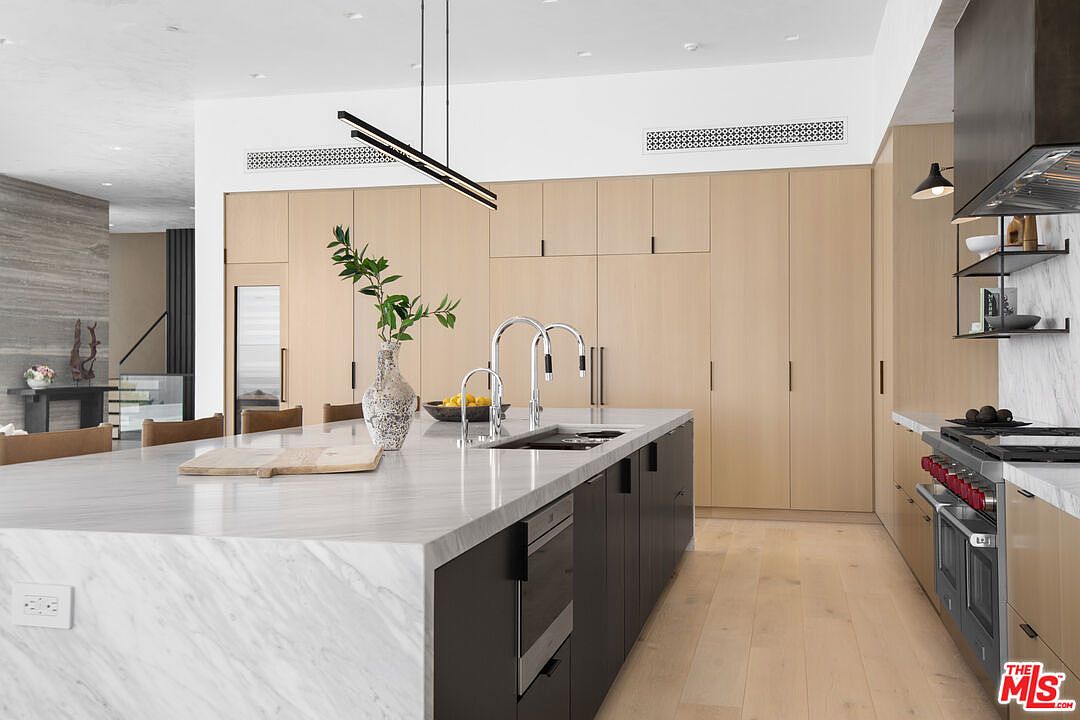This sophisticated, modern kitchen features a large marble-topped island with dark cabinetry and a sleek, minimalist aesthetic. The space is defined by floor-to-ceiling light wood cabinetry, high-end stainless steel appliances, and elegant pendant lighting. The perspective captures the clean lines and open-concept design, emphasizing a luxurious and functional culinary environment.