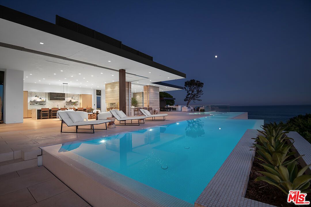 This stunning modern luxury estate features a seamless indoor-outdoor transition, highlighted by a sleek infinity-edge pool overlooking the ocean at twilight. The expansive patio is furnished with contemporary lounge chairs, while the open-concept interior reveals a sophisticated kitchen and living area illuminated by recessed lighting. The architectural design emphasizes clean lines, natural materials, and a breathtaking panoramic view of the coastline under a moonlit sky.