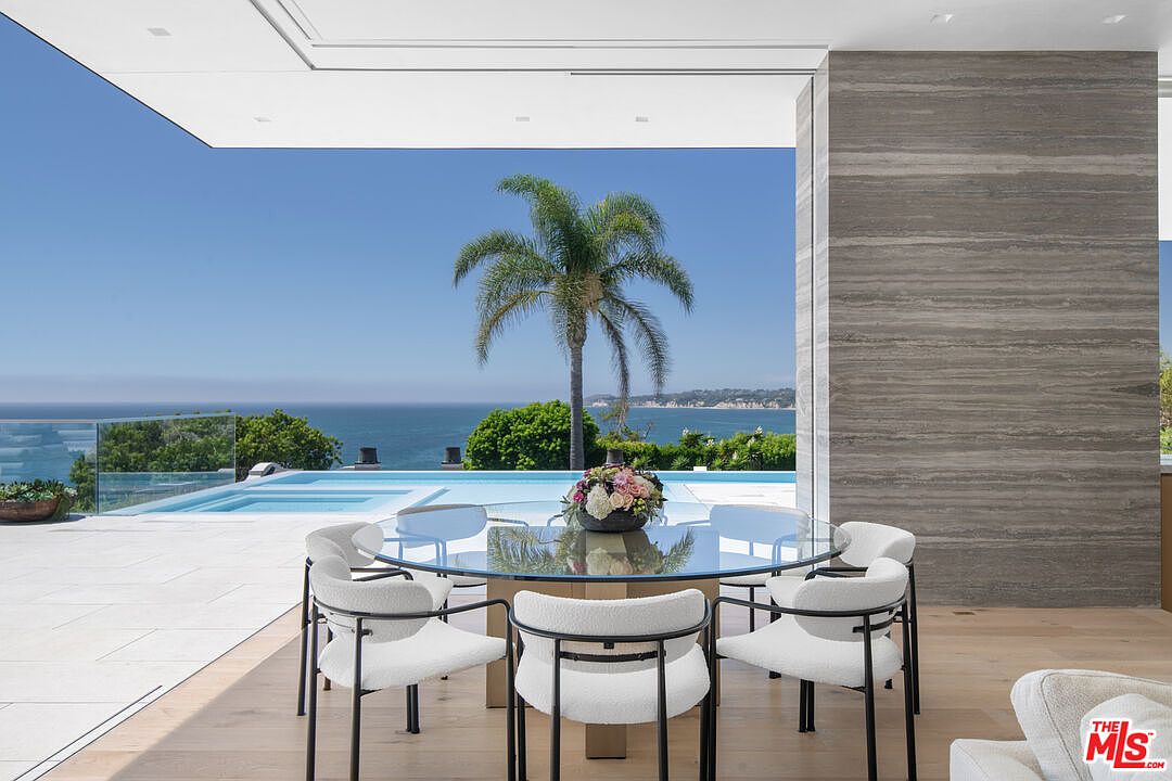 This elegant dining area features a sleek glass-topped table surrounded by modern, cream-colored upholstered chairs with black metal frames. The room seamlessly transitions to an expansive outdoor patio through floor-to-ceiling glass walls, showcasing a stunning infinity pool and a panoramic ocean view. The space is defined by a sophisticated blend of minimalist interior design and breathtaking natural scenery, creating a luxurious and serene atmosphere.