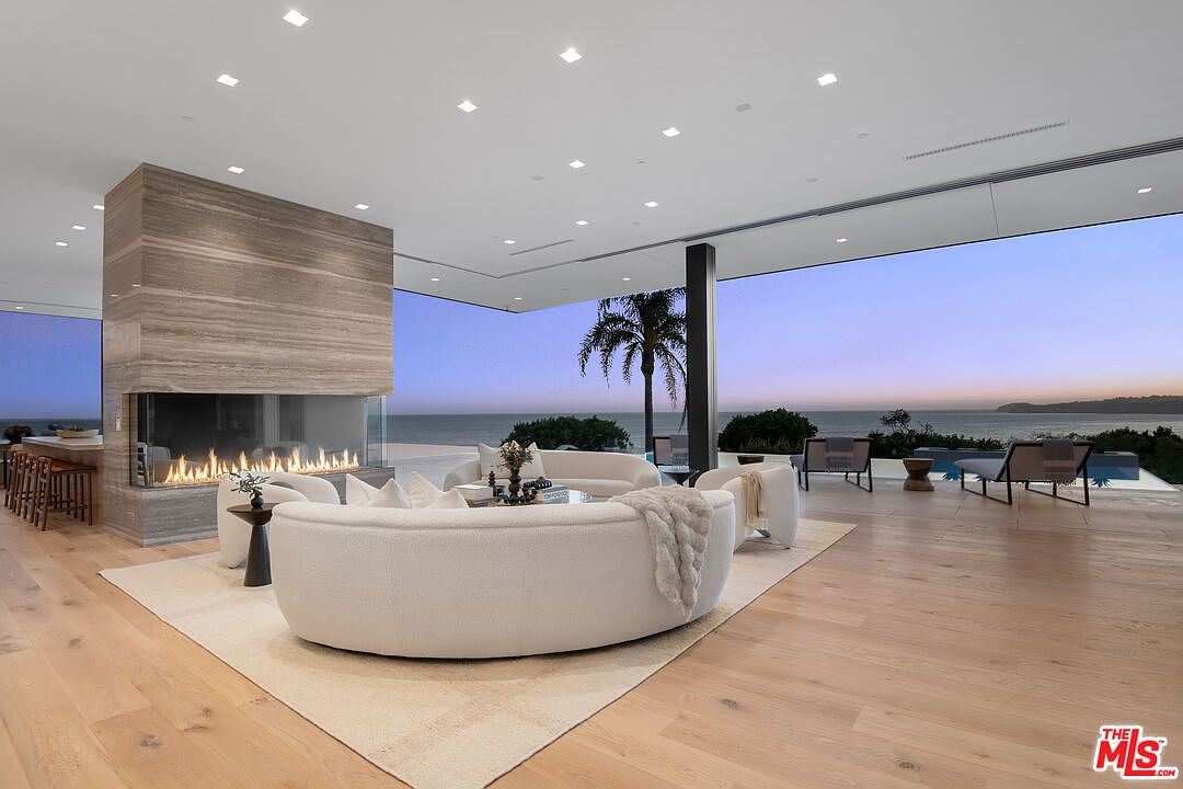 This expansive, ultra-modern living room features an open-concept design with floor-to-ceiling glass walls that seamlessly blend the interior with a breathtaking ocean view. A striking, large-scale stone fireplace serves as the central architectural anchor, complemented by plush, curved white sofas and light hardwood flooring. The space exudes a sophisticated, minimalist luxury, perfectly framing the serene coastal horizon at dusk.
