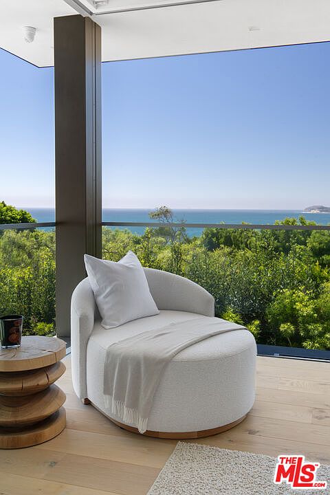 This serene balcony space features a plush, circular white lounge chair draped with a soft throw blanket, positioned to take full advantage of the panoramic ocean views. A rustic, stacked-wood side table sits beside the chair, grounding the modern, airy aesthetic of the space. The scene is framed by a sleek structural column and a glass railing, creating a seamless transition between the refined interior flooring and the lush greenery and blue horizon beyond.