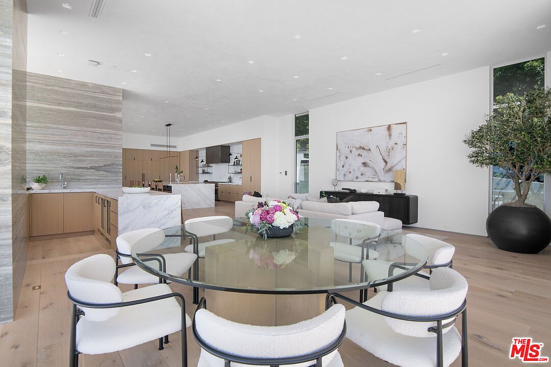 This expansive, open-concept living space features a contemporary dining area with a circular glass-top table surrounded by modern, cream-colored upholstered chairs. The room seamlessly transitions into a sleek, high-end kitchen with marble countertops and light wood cabinetry, while a comfortable living area with a plush sofa sits in the background. The bright, airy atmosphere is enhanced by minimalist decor, light hardwood flooring, and a large potted olive tree, creating a sophisticated and inviting modern aesthetic.