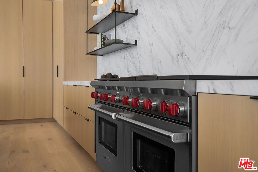 This close-up perspective highlights a high-end Wolf range with its signature red knobs, set against a sleek marble backsplash and light wood cabinetry. The minimalist design is complemented by open shelving above, creating a sophisticated and modern culinary space. The composition emphasizes the premium quality of the appliances and the clean, contemporary aesthetic of the kitchen.