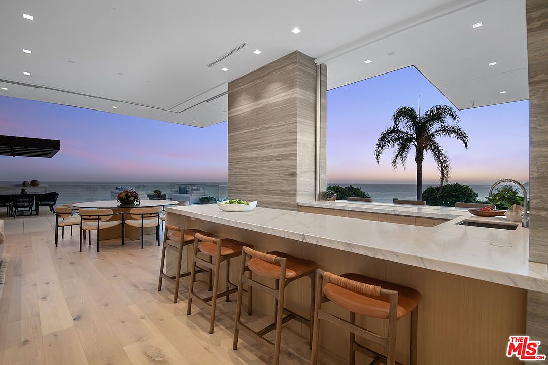 This sophisticated, open-concept kitchen features a massive marble-topped island with warm-toned wooden bar stools, seamlessly blending into a dining area. The space is defined by its floor-to-ceiling glass walls that offer an expansive, unobstructed view of the ocean and a palm tree at sunset. The minimalist design, light wood flooring, and recessed lighting create a luxurious, airy atmosphere perfect for high-end coastal living.