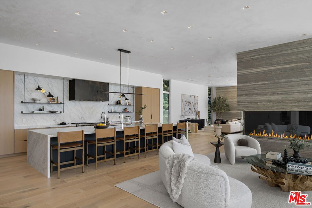 This expansive, open-concept kitchen and living area showcases a sophisticated modern aesthetic with high-end finishes, including a large marble-topped island and sleek dark cabinetry. The space is anchored by a striking floor-to-ceiling stone fireplace that seamlessly connects the kitchen to the adjacent living room, which features plush, curved seating. The bright, airy atmosphere is enhanced by recessed lighting and light-toned hardwood floors, creating a luxurious and inviting environment perfect for entertaining.