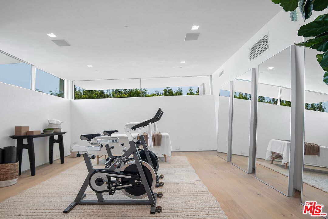This bright and minimalist home gym features a stationary exercise bike as the focal point, set upon a textured area rug on light wood flooring. The room is equipped with a massage or treatment table in the background and large, floor-to-ceiling mirrors that enhance the sense of space and light. High, clean white walls and clerestory windows create an airy, serene atmosphere perfect for a wellness or fitness retreat.