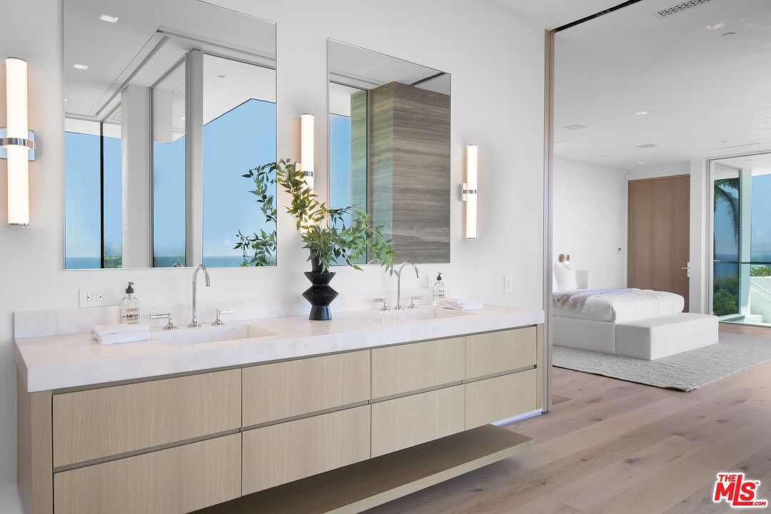 This luxurious primary bathroom features a sleek, double-vanity with light wood cabinetry and a pristine white stone countertop. Two large, frameless mirrors are mounted above the sinks, reflecting the bright, airy space and the adjacent bedroom suite. The minimalist design, combined with high-end fixtures and a view of the ocean through the bedroom window, creates a sophisticated and tranquil atmosphere.