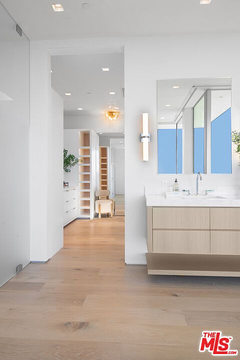 This modern primary bathroom features a sleek, floating light-wood vanity with a white countertop and a rectangular mirror, illuminated by a contemporary wall sconce. The space flows seamlessly into a bright, minimalist walk-in closet area with light hardwood flooring throughout. The clean lines, neutral color palette, and recessed lighting create an airy, sophisticated atmosphere.