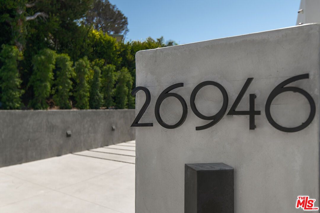 A close-up, eye-level shot focuses on a modern concrete pillar displaying the house number '26946' in sleek, black, sans-serif metal numerals. The background features a blurred view of a paved driveway and a lush, manicured hedge, suggesting a contemporary and private residential setting. The clean lines and minimalist aesthetic convey a sense of modern luxury and architectural precision.