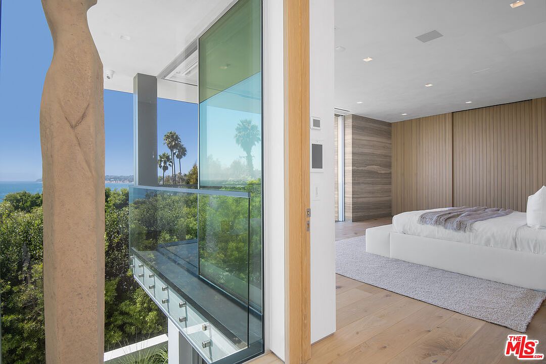 This sophisticated primary bedroom features a seamless transition to a private balcony through large glass sliding doors, offering breathtaking ocean and treetop views. The room is designed with a minimalist aesthetic, showcasing warm wood-paneled walls, light hardwood flooring, and a plush white bed. The perspective captures the elegant interplay between the serene indoor sanctuary and the expansive, sun-drenched outdoor landscape.