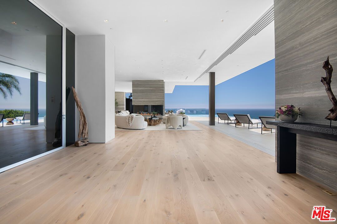 This expansive, ultra-modern living room features light-toned hardwood flooring and seamless indoor-outdoor flow through floor-to-ceiling glass walls that open to a panoramic ocean view. The space is anchored by a minimalist stone fireplace and contemporary white furniture, creating an airy, sophisticated atmosphere. The perspective captures the transition from the interior living area to the sun-drenched patio and infinity pool beyond.