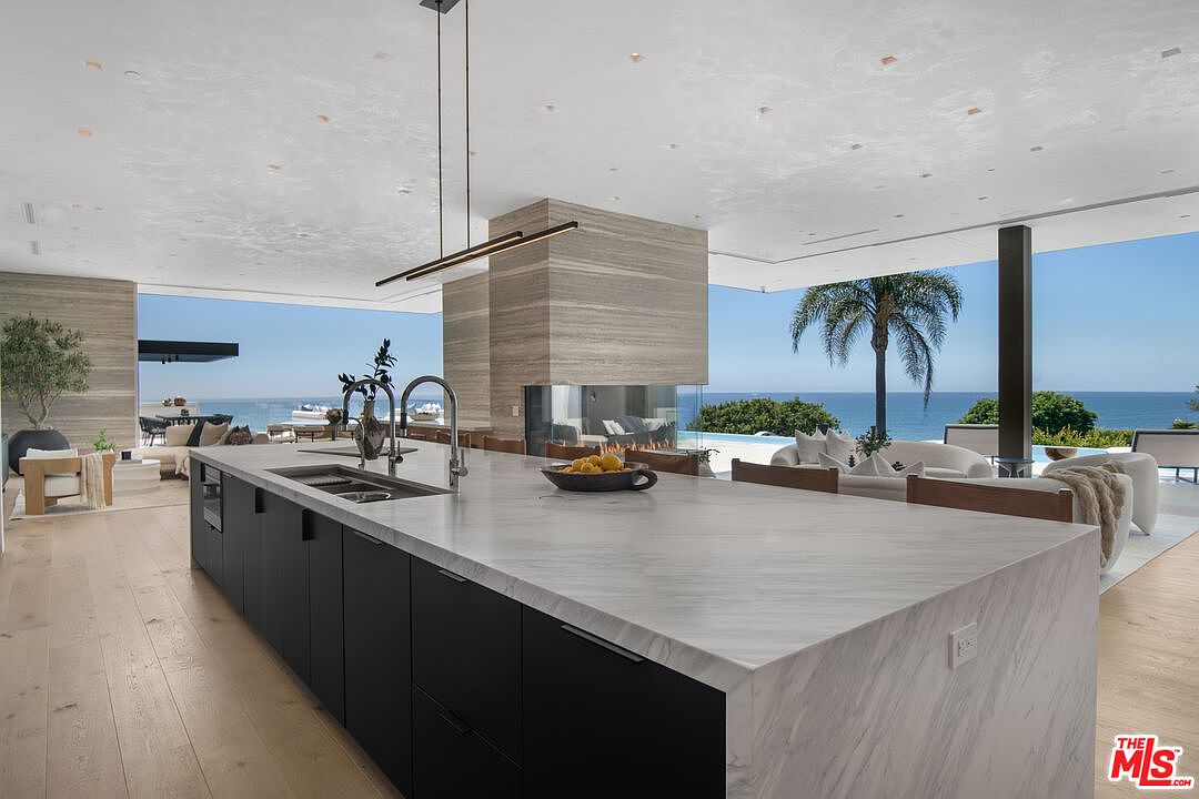 This ultra-modern, open-concept kitchen features a massive waterfall-edge marble island with matte black cabinetry, serving as the centerpiece of a luxurious living space. The room seamlessly transitions to an expansive outdoor patio and pool area through floor-to-ceiling glass walls, offering breathtaking, unobstructed ocean views. The design is defined by its minimalist aesthetic, high-end finishes, and a striking stone-clad fireplace column that anchors the sophisticated, airy atmosphere.