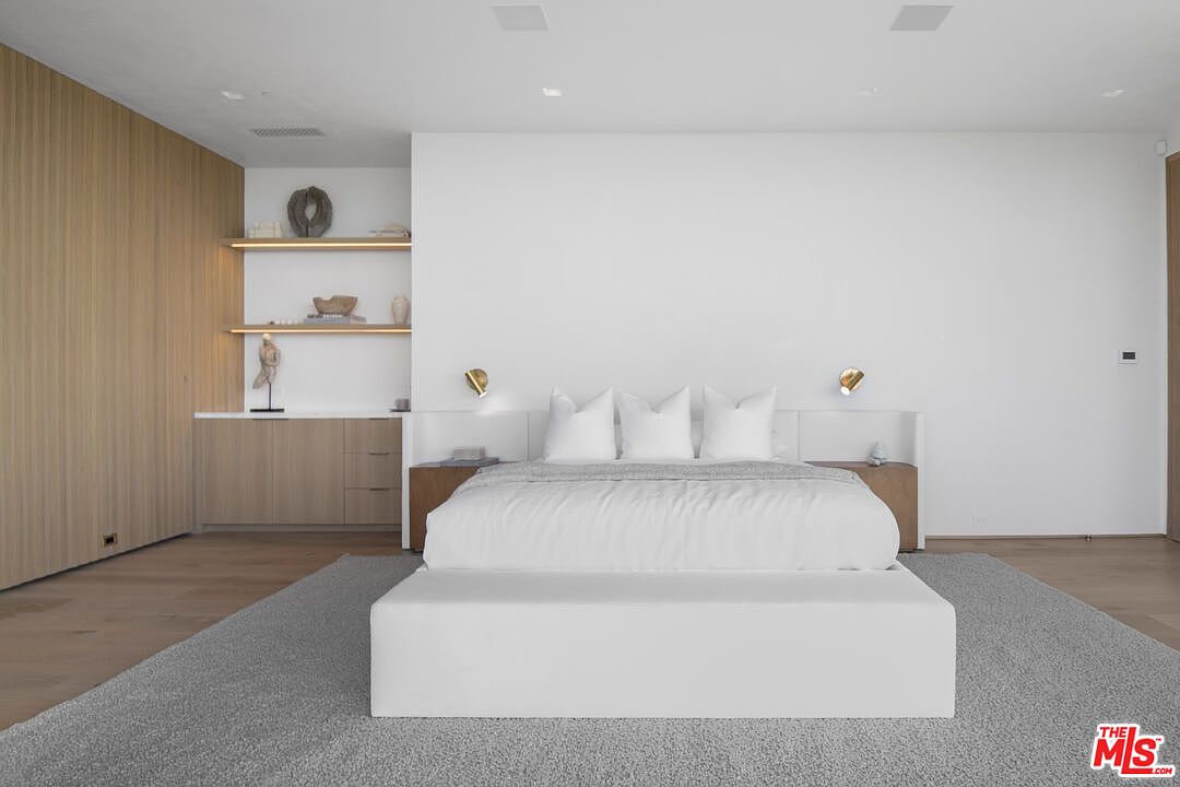 This serene primary bedroom features a minimalist, contemporary design with a neutral color palette and warm wood accents. The room is anchored by a plush white bed centered against a clean white wall, flanked by integrated wooden nightstands and sleek wall-mounted reading lights. To the left, a built-in shelving unit with recessed lighting adds a sophisticated touch, while a large, textured area rug provides comfort and warmth to the space.
