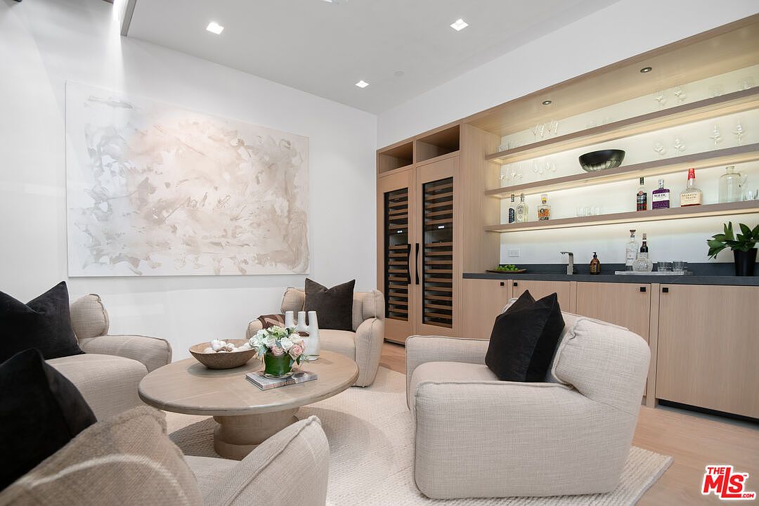 This sophisticated lounge area features a neutral color palette with plush, cream-colored armchairs arranged around a round wooden coffee table. A built-in wet bar with light wood cabinetry, open shelving, and a wine refrigerator adds a luxurious touch, while a large abstract painting serves as a focal point on the wall. The space exudes a calm, high-end atmosphere, perfect for entertaining or relaxing in a modern home.