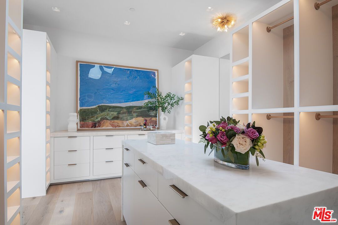 This luxurious walk-in closet features a central marble-topped island, custom white cabinetry with integrated lighting, and light wood flooring. A large, vibrant abstract painting serves as a focal point above the built-in dresser, while a sophisticated floral arrangement adds a touch of elegance to the space. The room is bright and airy, showcasing a high-end, organized design perfect for a primary suite.