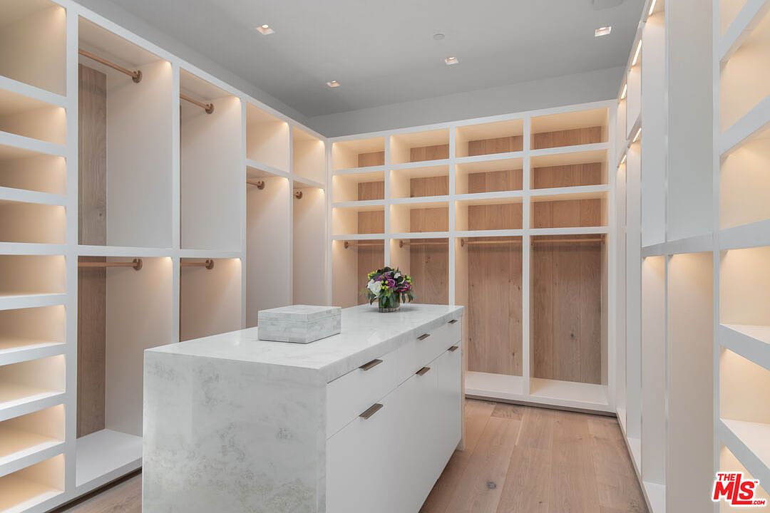 This luxurious, expansive walk-in closet features custom white cabinetry with integrated warm LED lighting and light wood-toned back panels. A central island topped with a white marble countertop provides additional storage and a sophisticated focal point, complemented by a small floral arrangement. The space is designed with a clean, modern aesthetic, emphasizing organization and high-end finishes.