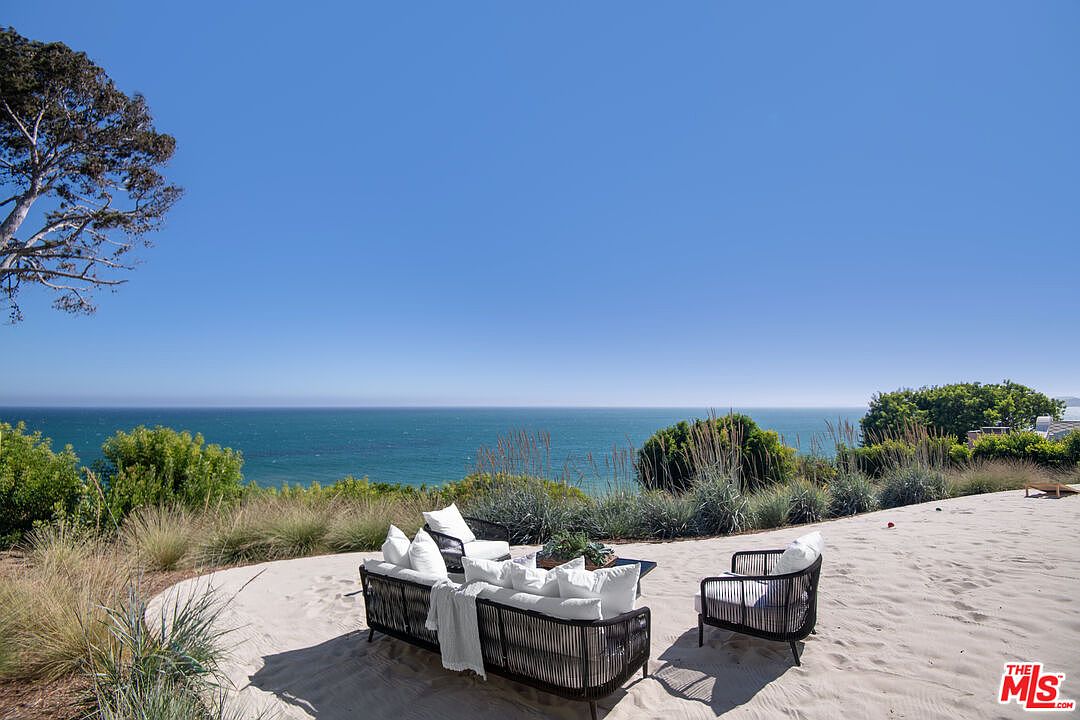 This stunning outdoor patio area features a modern, dark-wicker sectional sofa and matching armchair arranged on a sandy surface, perfectly positioned to overlook the vast, blue ocean. The scene is framed by native coastal grasses and a clear, cloudless sky, creating a serene and luxurious atmosphere. The perspective is elevated, emphasizing the seamless integration between the private living space and the expansive coastal horizon.