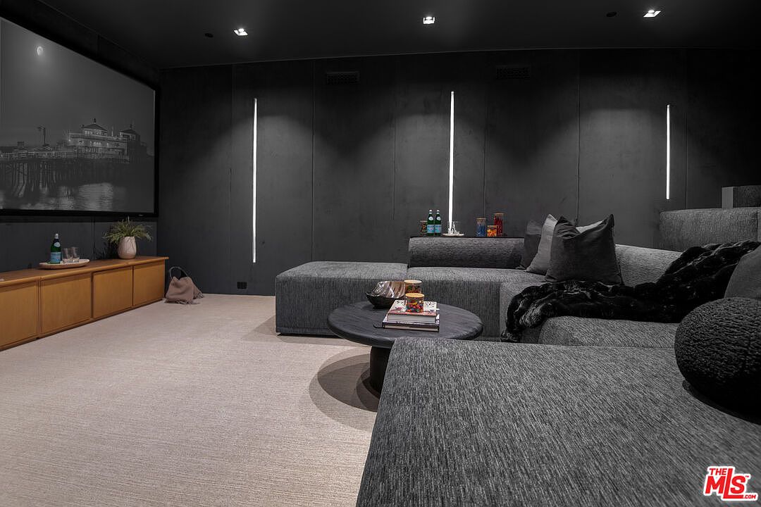 This sophisticated home theater features a large projection screen, plush charcoal-colored sectional seating, and a low-profile circular coffee table. The room is accented by sleek vertical LED light strips against dark, textured walls, creating an immersive and moody cinematic atmosphere. The perspective is from the foreground, looking toward the screen, emphasizing the comfortable and modern design of this dedicated entertainment space.