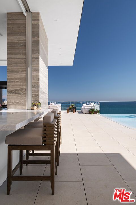 This luxurious outdoor terrace features a sleek, modern bar area with wooden stools overlooking a stunning infinity pool and the vast ocean horizon. The space is defined by clean lines, light-toned stone flooring, and a minimalist aesthetic that seamlessly blends indoor and outdoor living. The perspective captures the serene atmosphere of a high-end coastal property, emphasizing the expansive views and sophisticated design.
