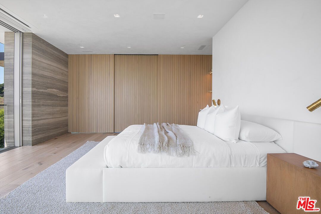 This luxurious primary bedroom features a minimalist, contemporary design with a low-profile white platform bed as the central focal point. The room is accented by a striking floor-to-ceiling wood-paneled wall and a large sliding glass door that opens to an outdoor balcony, bathing the space in natural light. The neutral color palette, soft area rug, and clean lines create a serene and sophisticated atmosphere.